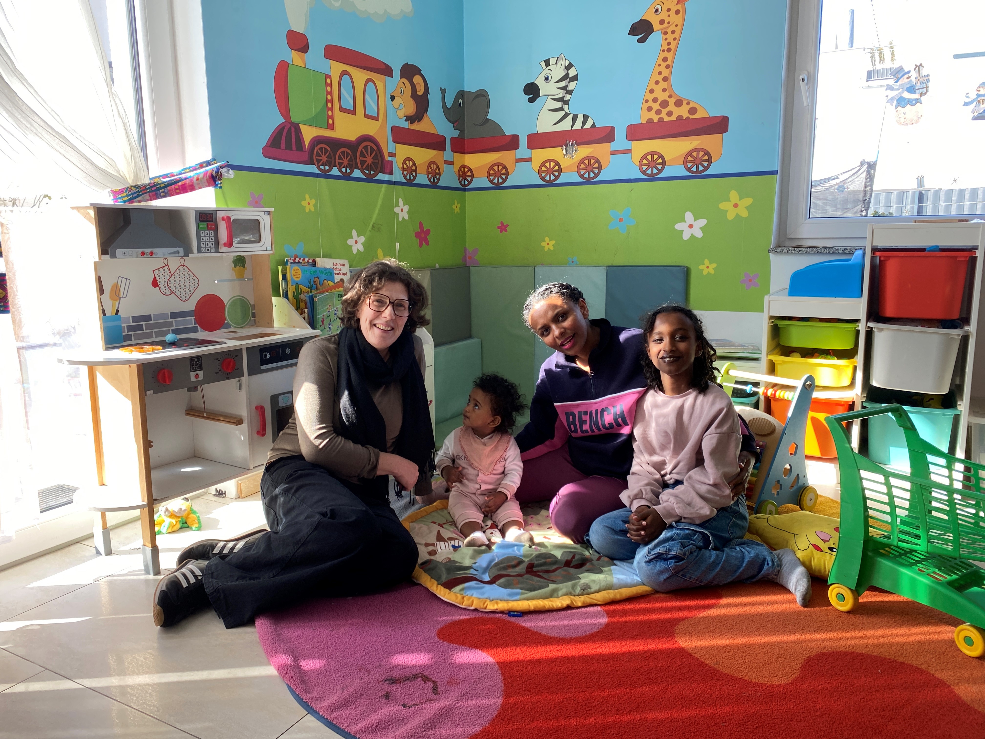 Kathrin Smola-Peter vom Fachdienst Kinderbetreuung zusammen mit Heaven Zewdu-Tekeste und ihren Töchtern Abigia und Delina auf dem Boden sitzend in einer bunten Spielecke
