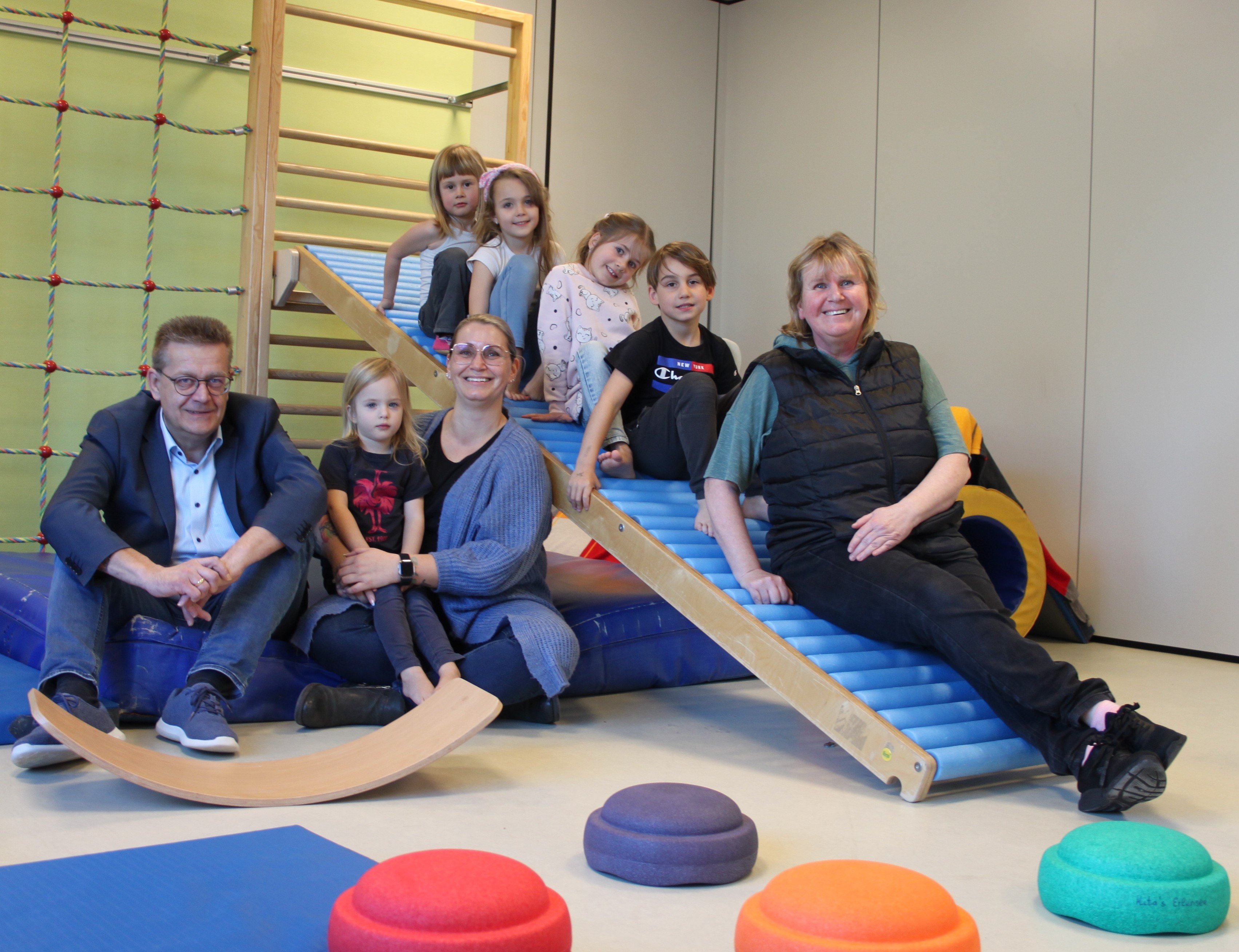Bürgermeister Stefan Erb und Kita-Leiterin Nina Muth besuchen Ellen Krause im Turnraum. Sie sitzen mit einigen KiTa-Kindern auf Turngräten und Matten.