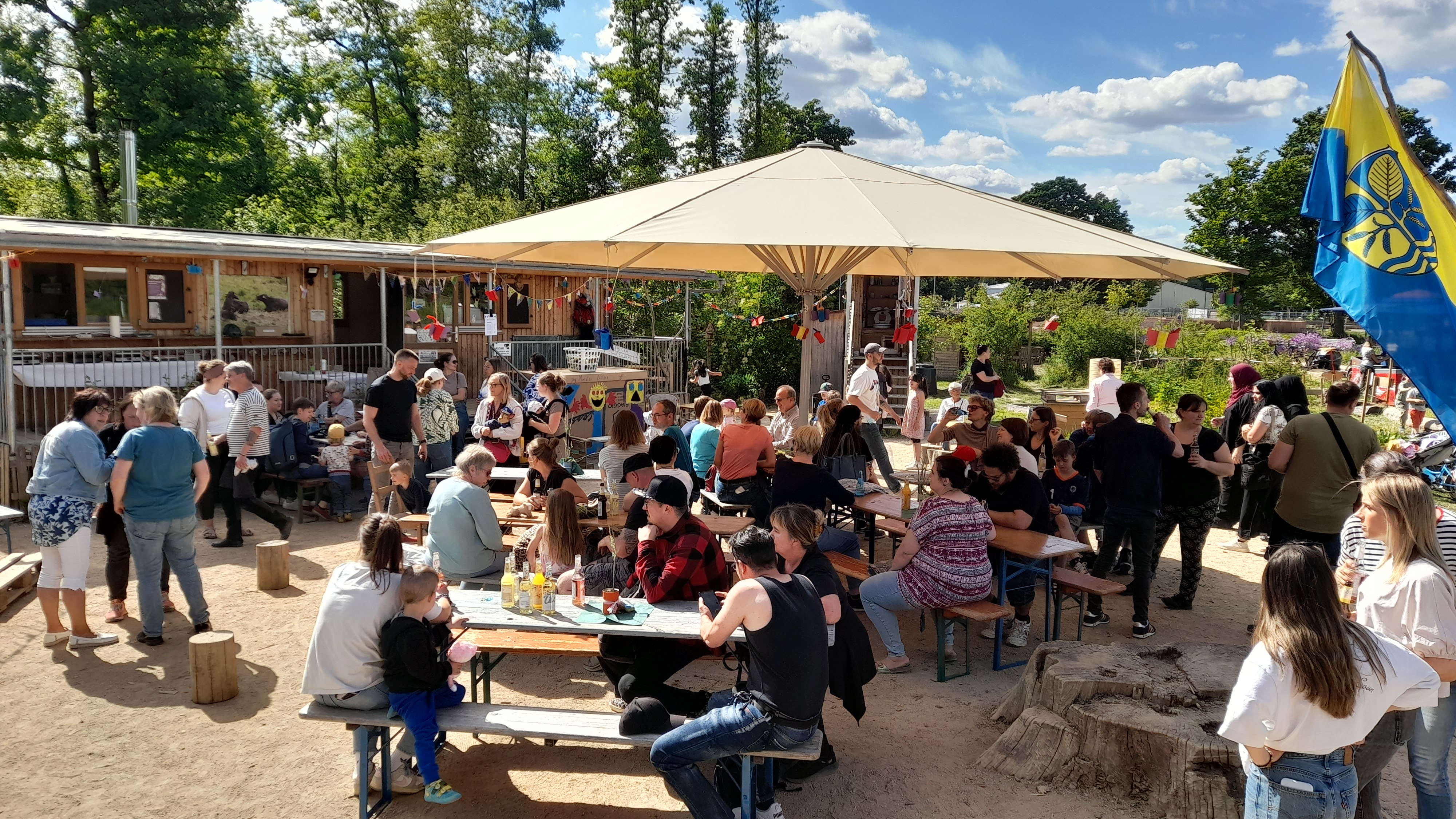 Viele Menschen sitzen an Tischen unter einem Sonnenschirm vor einem Bauwagen