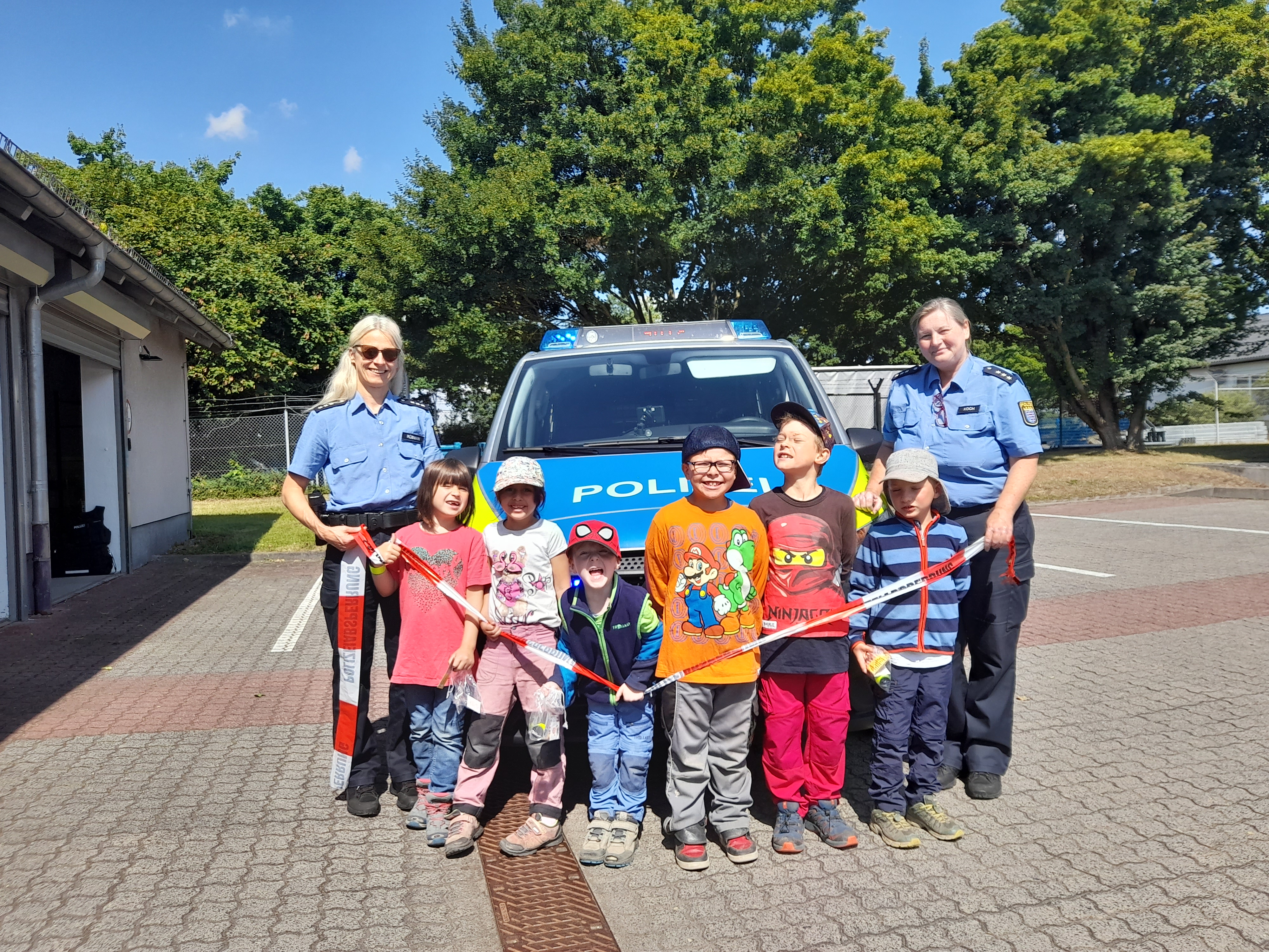 KiTa-Kinder mit zwei Polizeibeamtinnen stehen vor einem Polizeiauto