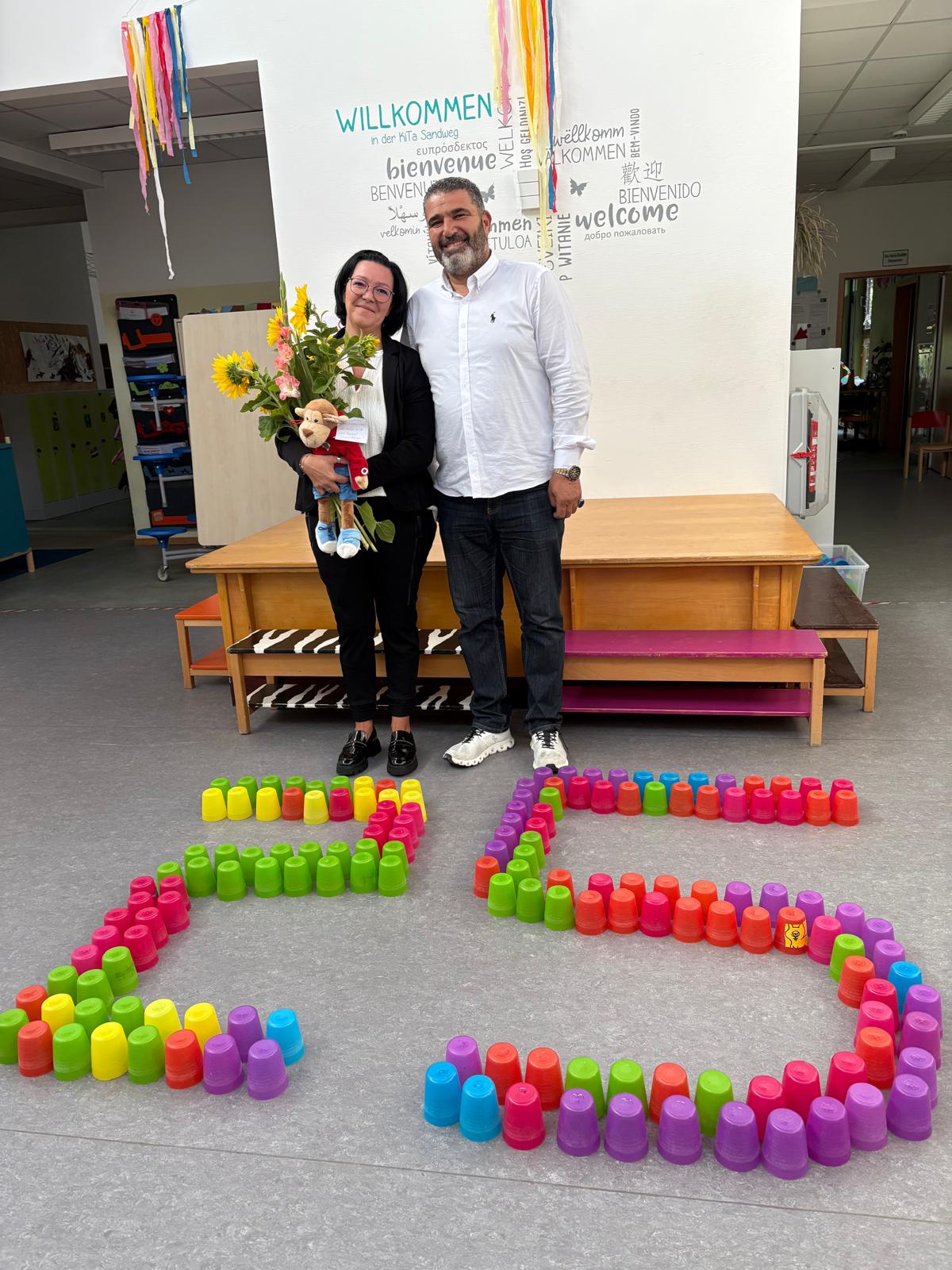 Jane Matzke und mit Erzieher Emad Nayleh vor einer aus bunten Bechern gestellten 25