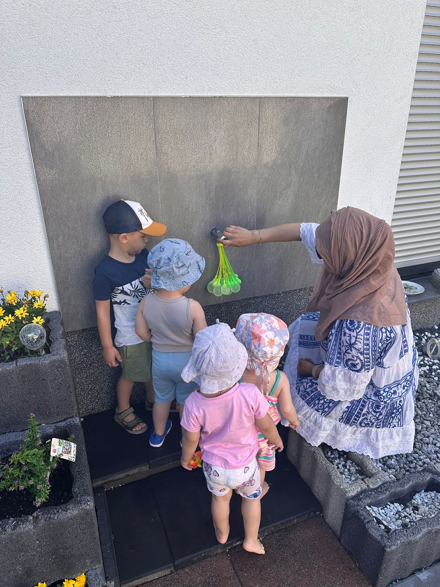 Kinder füllen kleine Wasserballons am Wasserhahn im Garten mit Ihrer Tagesmutter