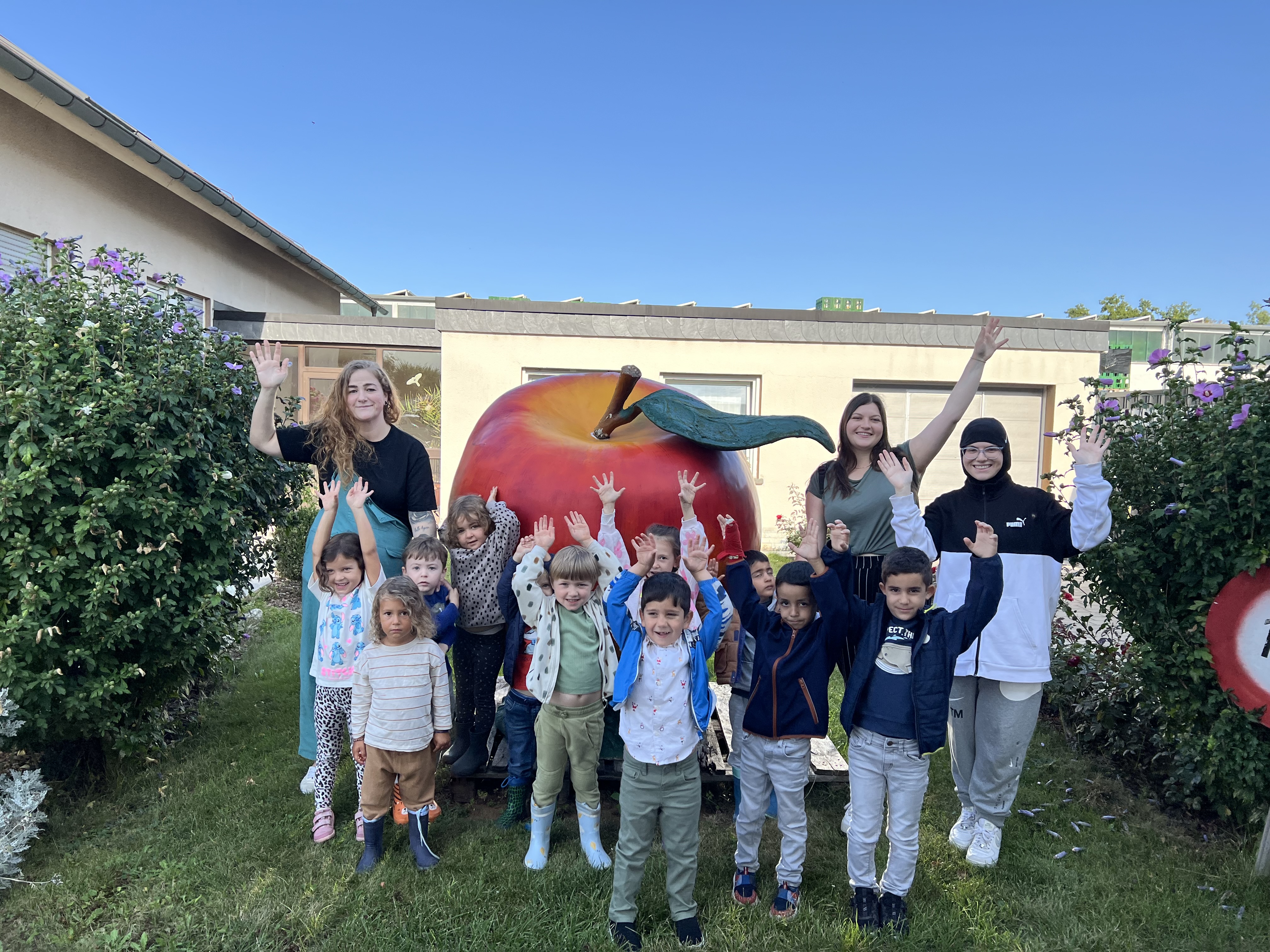 KiTa-Kinder und Erzieherinnen winken vor einem großen Apfelmodell