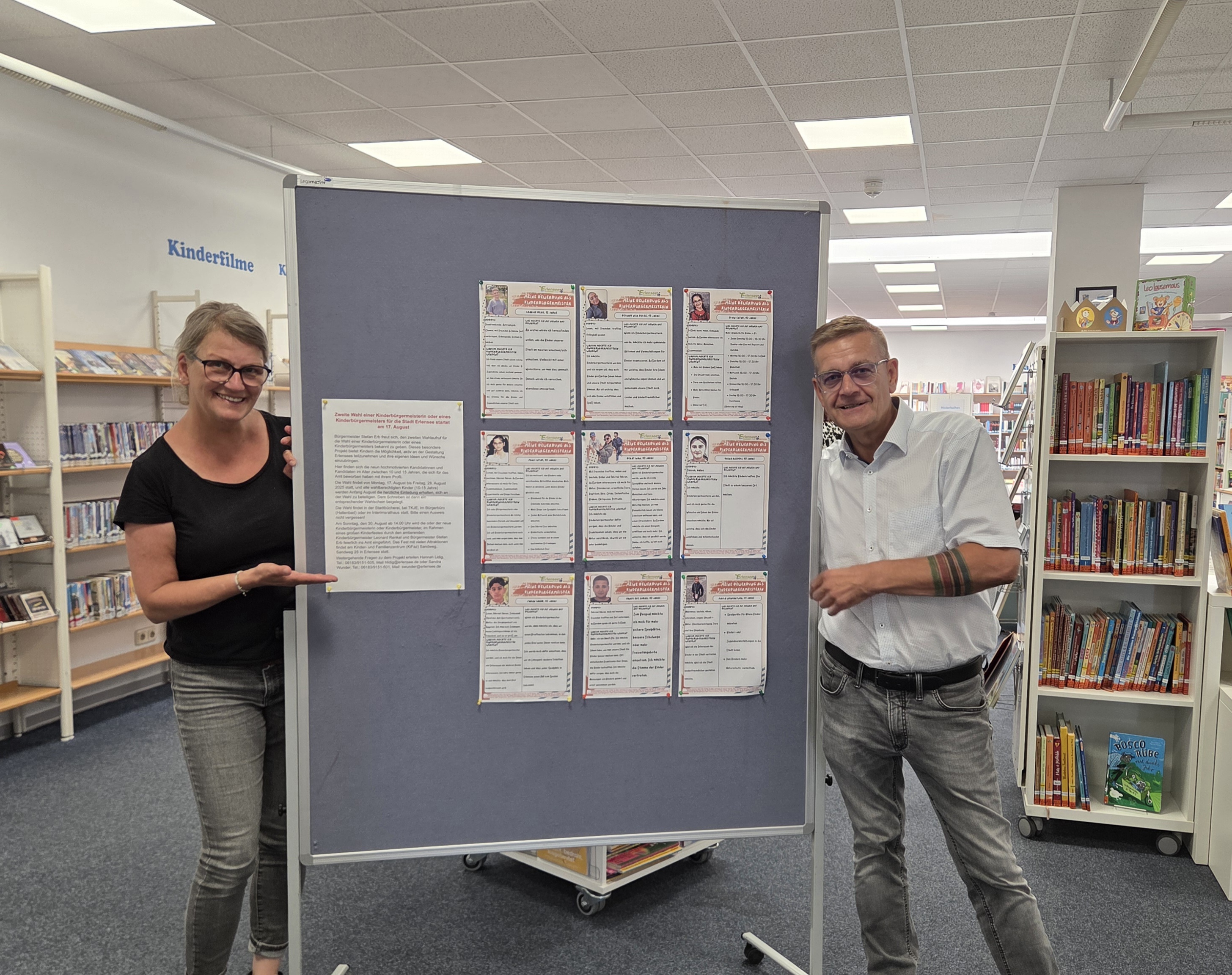 Bürgermeister Stefan Erb und Fachdienstleitung Sandra Wunder stehen bei einer Pinnwand, an welcher die Profile der Kandidatinnen und Kandidaten zur Kinderbürgermeisterin oder zum Kinderbürgermeister ausgehängt sind.