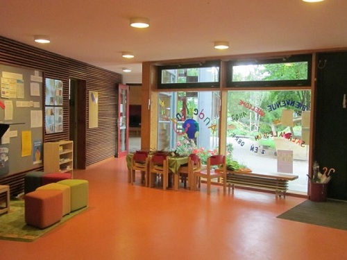 Foyer der Evangelischen Kita Anderland. Bodentiefe Fenster zum Aussenbereich. Davor eine Stuhlecke und eine Bank. Orangener Boden. Links ist eine weitere Sitzecke mit Hockern und einer Pinwand zu sehen.