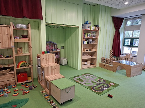 Bauraum der evangelischen Kita Anderland. Der Boden und die holzvertaefelten Waende sind mintgruen. Im Raum sind viele verschiedene Holzbausteine, ein Verkehrsteppich und kleine Fahrzeuge und Figuren auf dem Boden und in den verschiedenen Regalen zu sehen. Auf der rechten Seite sind Fenster mit Blick zum Aussengelaende.