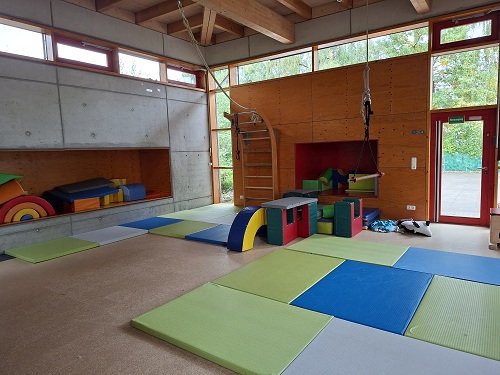 Bewegungsraum der evangelischen Kita Anderland. Auf dem Boden liegen Matten in gruen, blau und grau. Zudem sind Schaumstoffwuerfel in verschiedenen Formen verteilt. An der Wand befindet sich eine Kletterwand mit Ueberhang. Von der Decke haengt ein Kletterseil und eine Haengestange. Oberhalb des hohen Raumes befinden sich ringsum Fenster. Die Seitenfenster und die Glastuer blicken nach draussen.