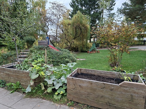 Garten der evangelischen Kita Anderland. Im vorderen Bereich sind zwei Hochbeete mit verschiedenen Gemuesepflanzen zu sehen. Im Hintergrund ist links eine rote Rutsche in einen Erdhuegel eingebaut zu sehen, die auf eine Wiese mit verschiedener Bepflanzung fuehrt.