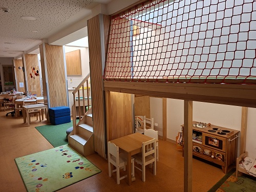 Spielflur der Evangelischen Kita Anderland. Auf der rechten Seite führen Treppen nach oben zu einer Zwischenebene. Diese ist mit einem Netz gesichert. Auf der Unteren Ebene ist eine Spielkueche und ein kleiner Tisch mit Stuehlen zu sehen.
